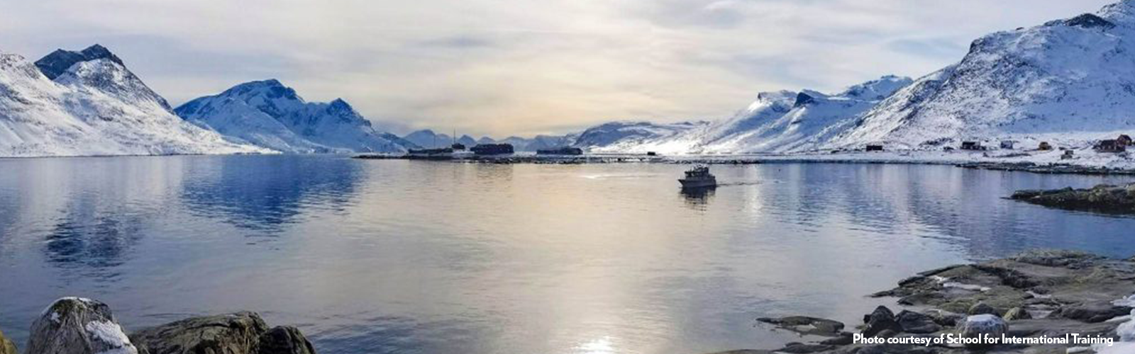 The sun reflecting over a lake surrounded by snowy mountains.